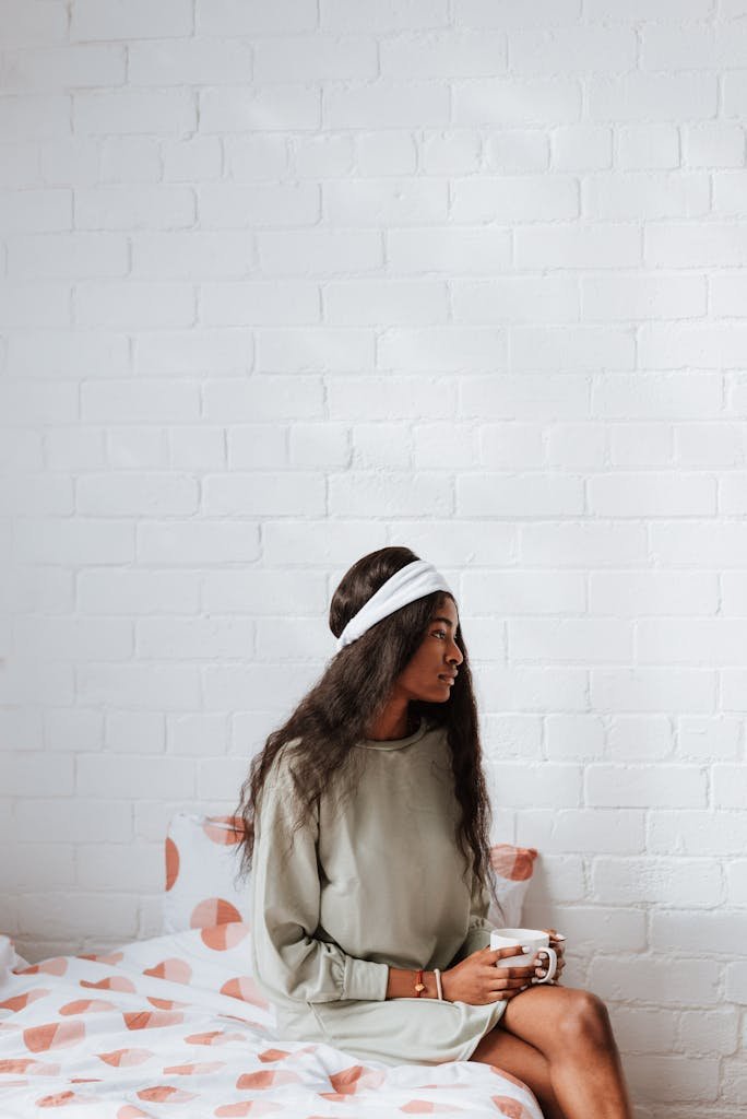 A young woman with long hair sits on a bed, holding a mug, in a bright, minimalist bedroom.